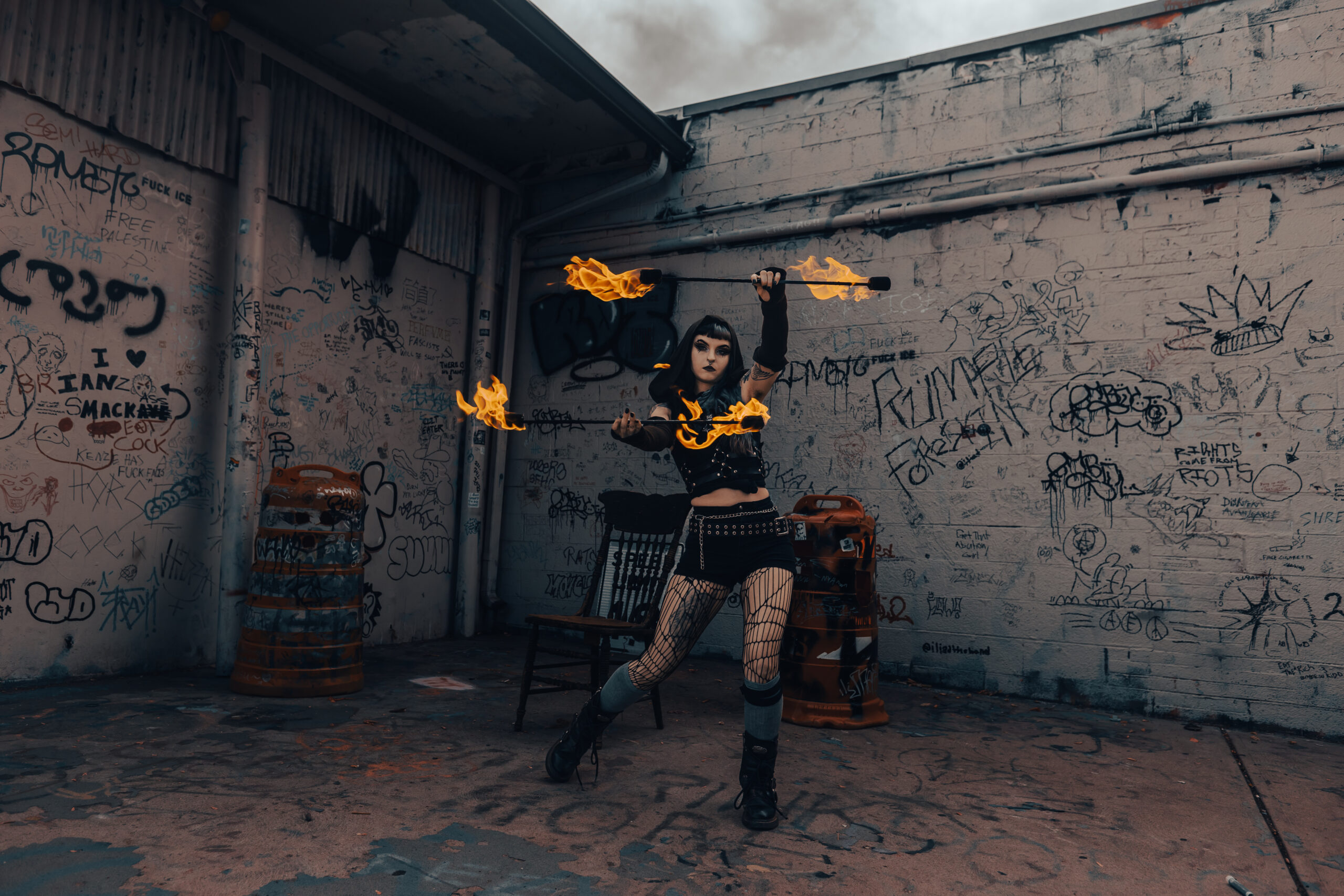 Performer spins flaming batons in a graffiti-covered alley.