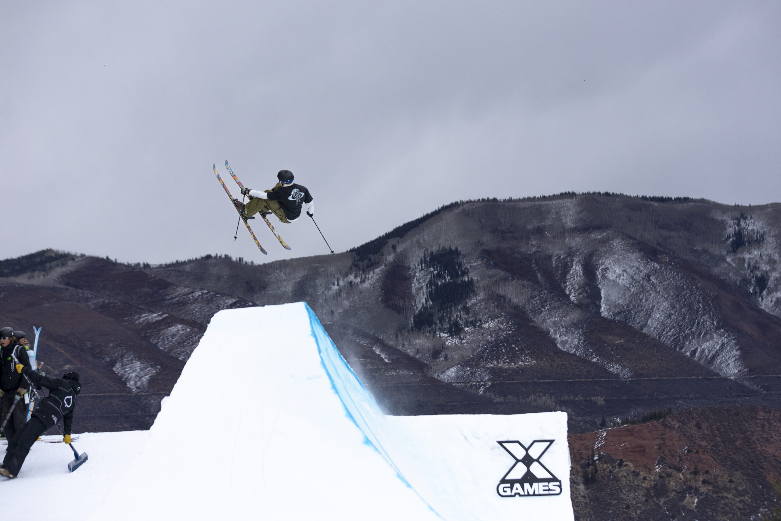 Freeskier launches above an X Games jump with mountains behind.